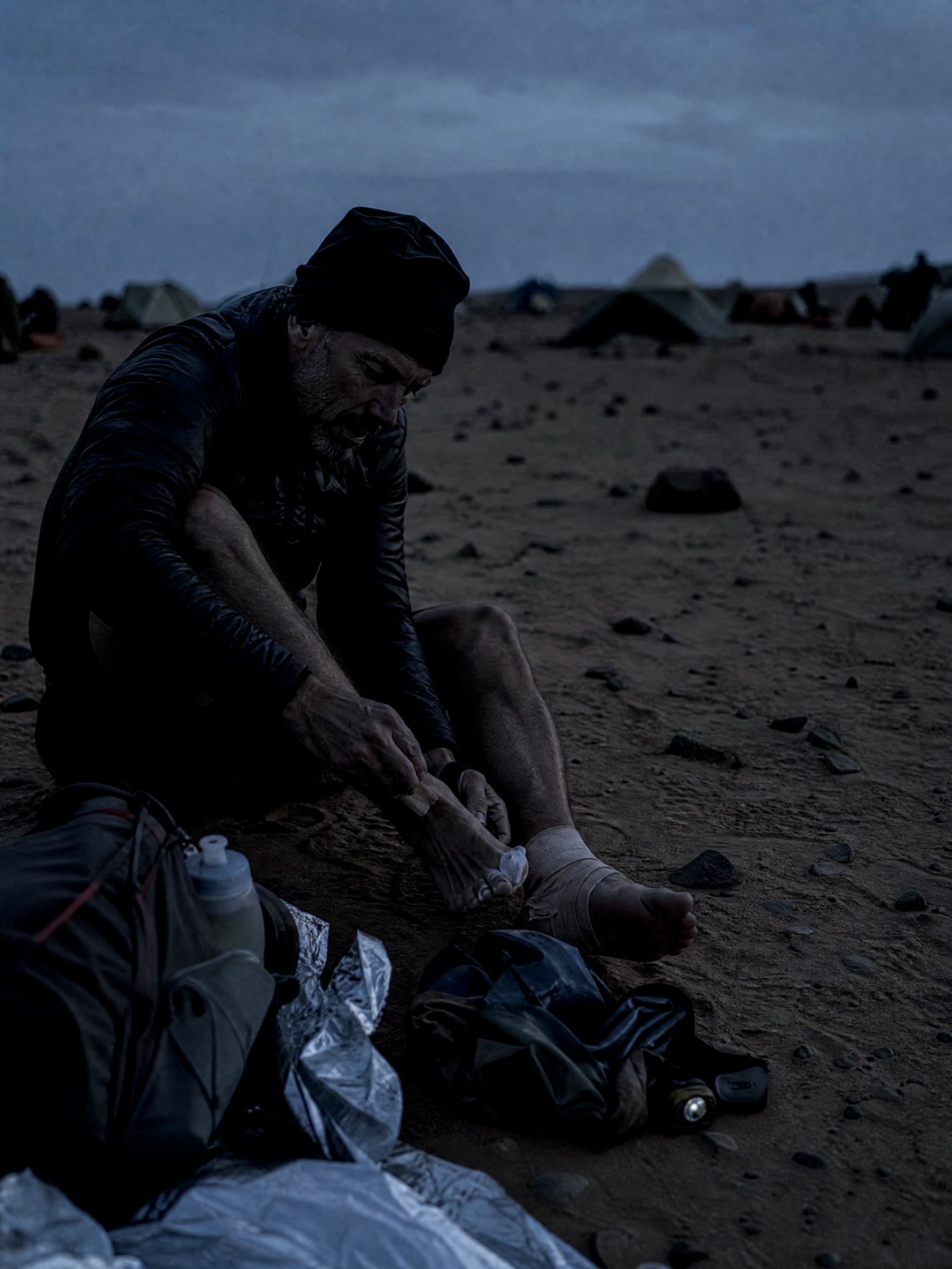 A runner taping his feet at dusk in a desert bivouac, tents and headtorch beside him