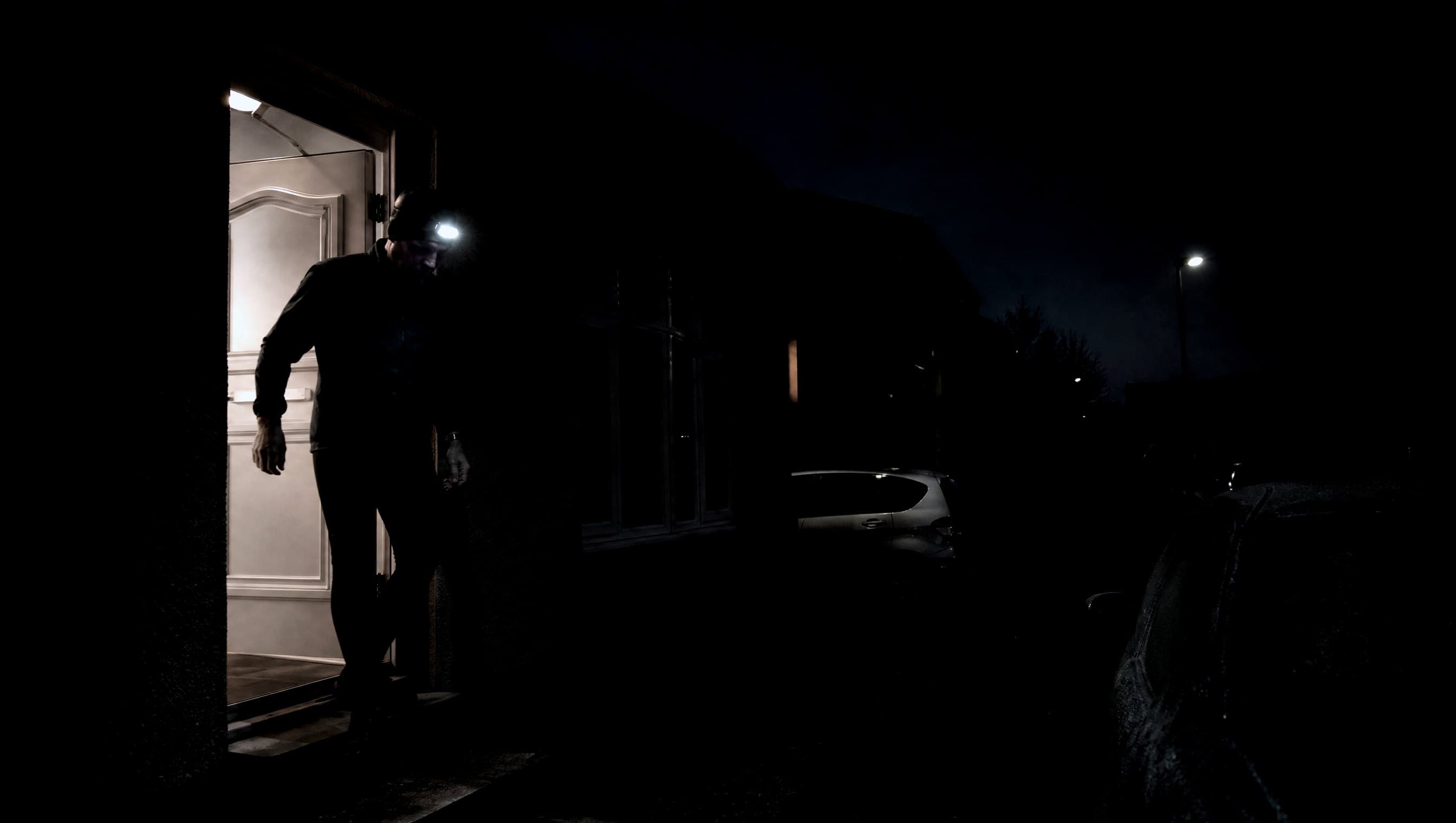 A masters-age runner leaving a suburban front door before dawn with a headtorch on, frost on the parked cars