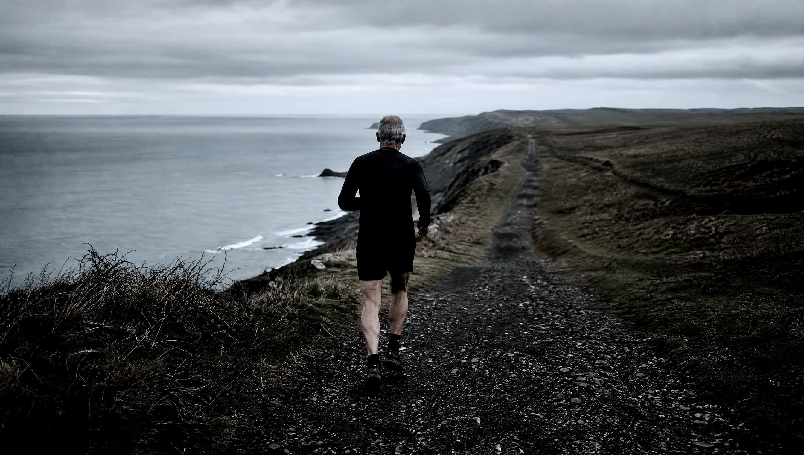 A masters-age runner seen from behind on a long empty clifftop path under flat grey light