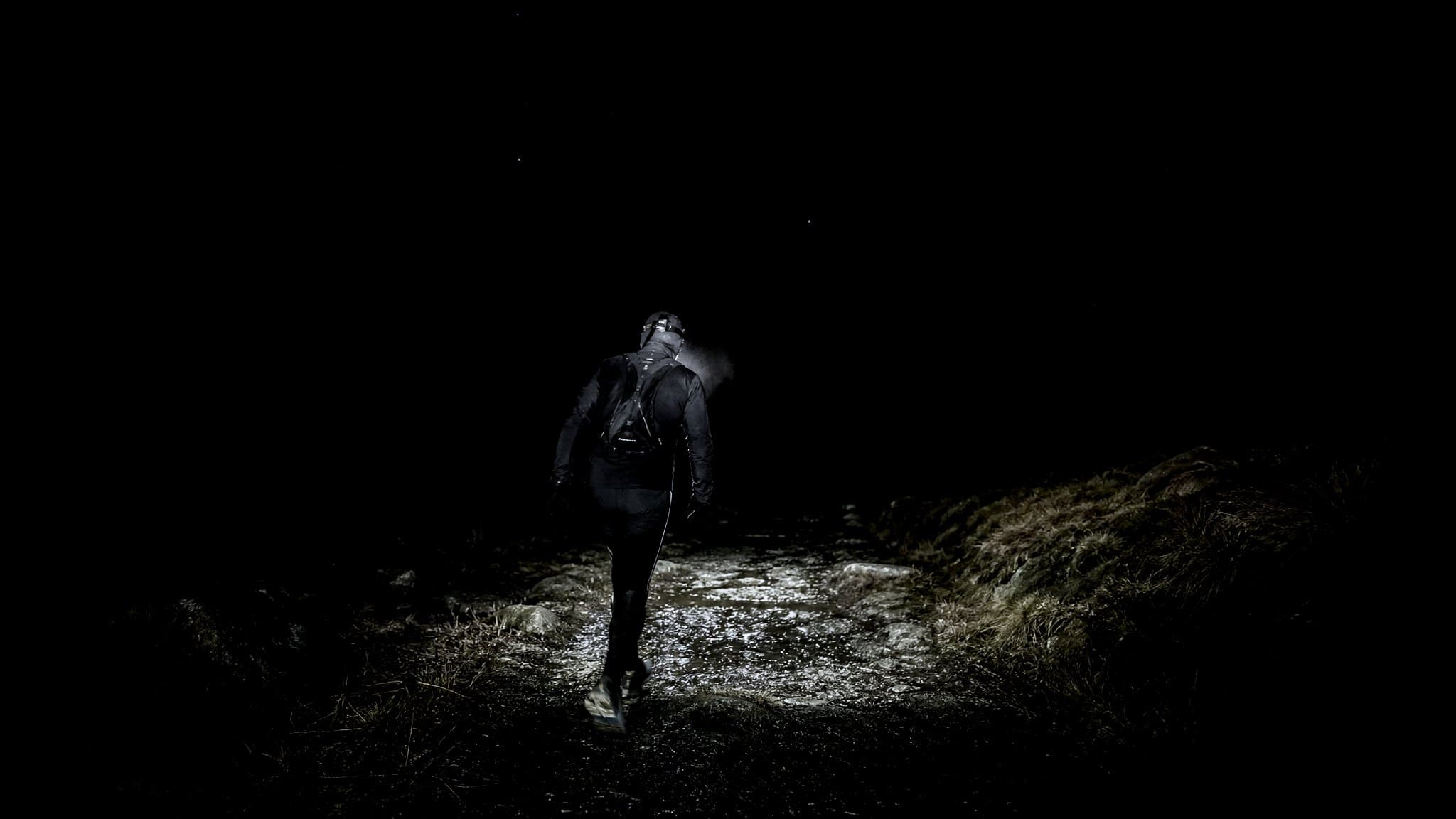 A masters-age ultra runner alone on a moorland track at night, headtorch lighting the wet stony path ahead