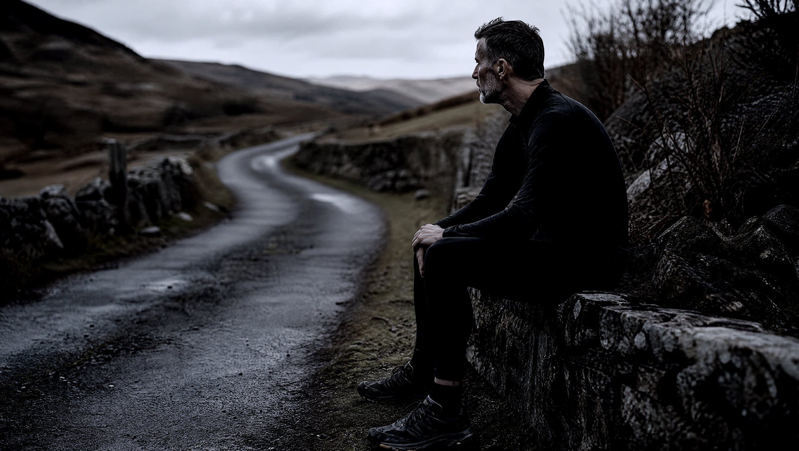A masters-age runner sitting still on a stone wall beside a quiet lane in early morning light