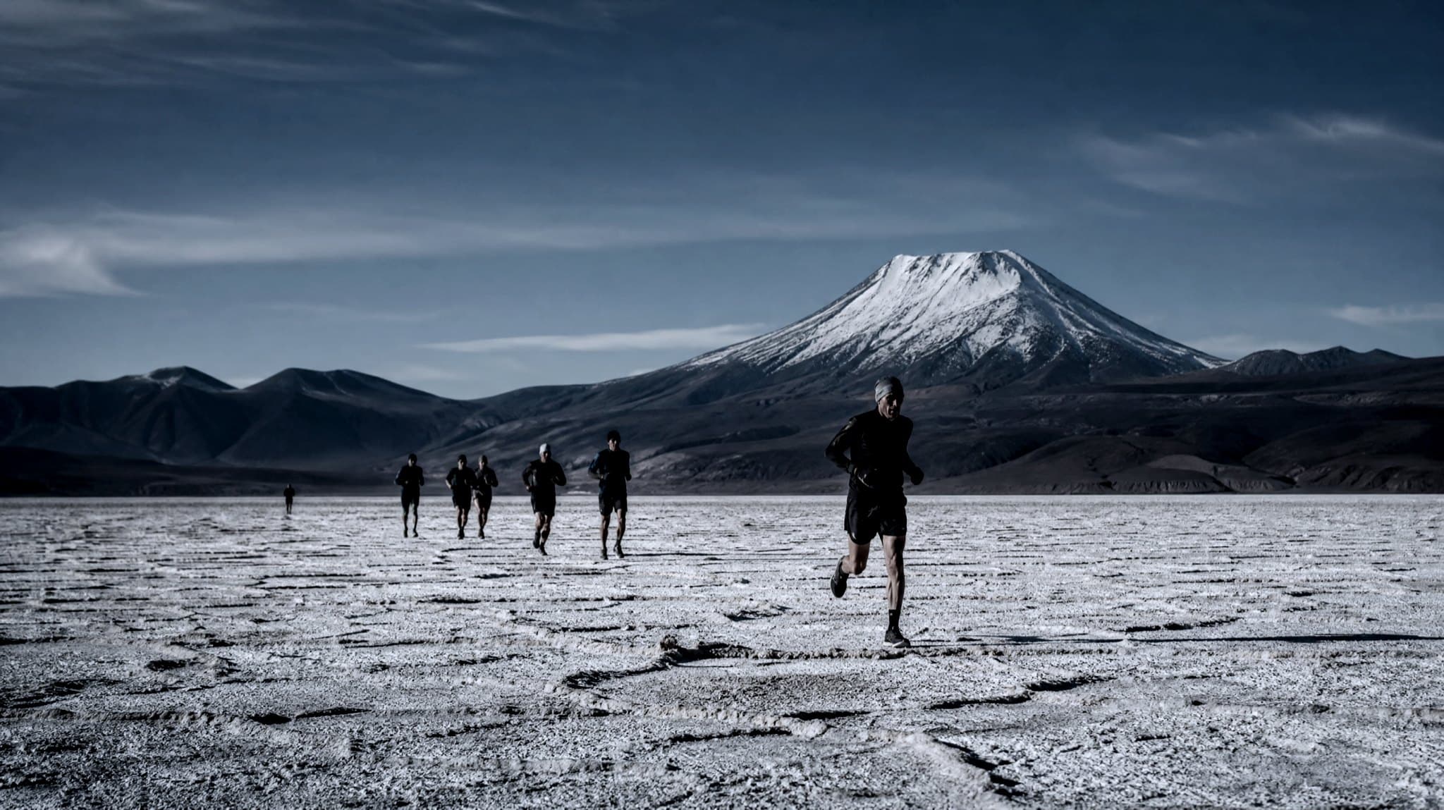 A masters-age runner with a loaded pack crossing a cracked white salt flat with volcanoes on the horizon
