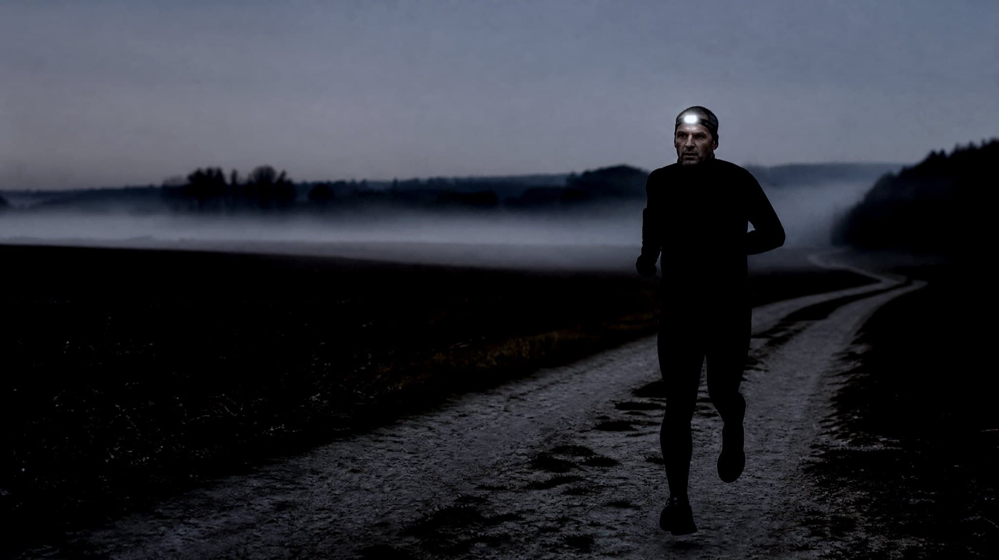 A masters-age runner with a head torch on a misty track at dusk in autumn