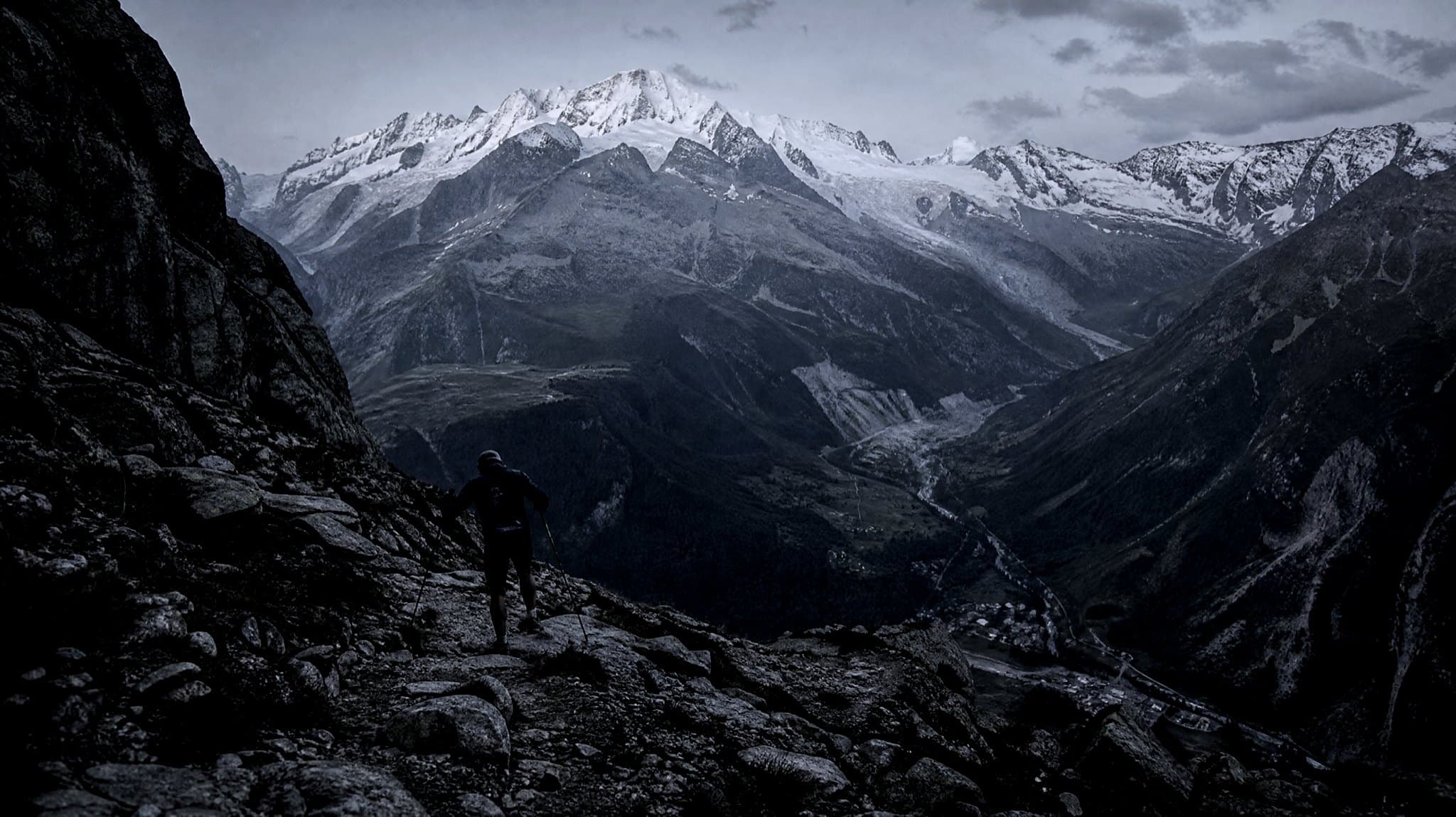 A masters-age runner with poles descending a rocky alpine path at dusk, snow-covered peaks and a deep valley ahead