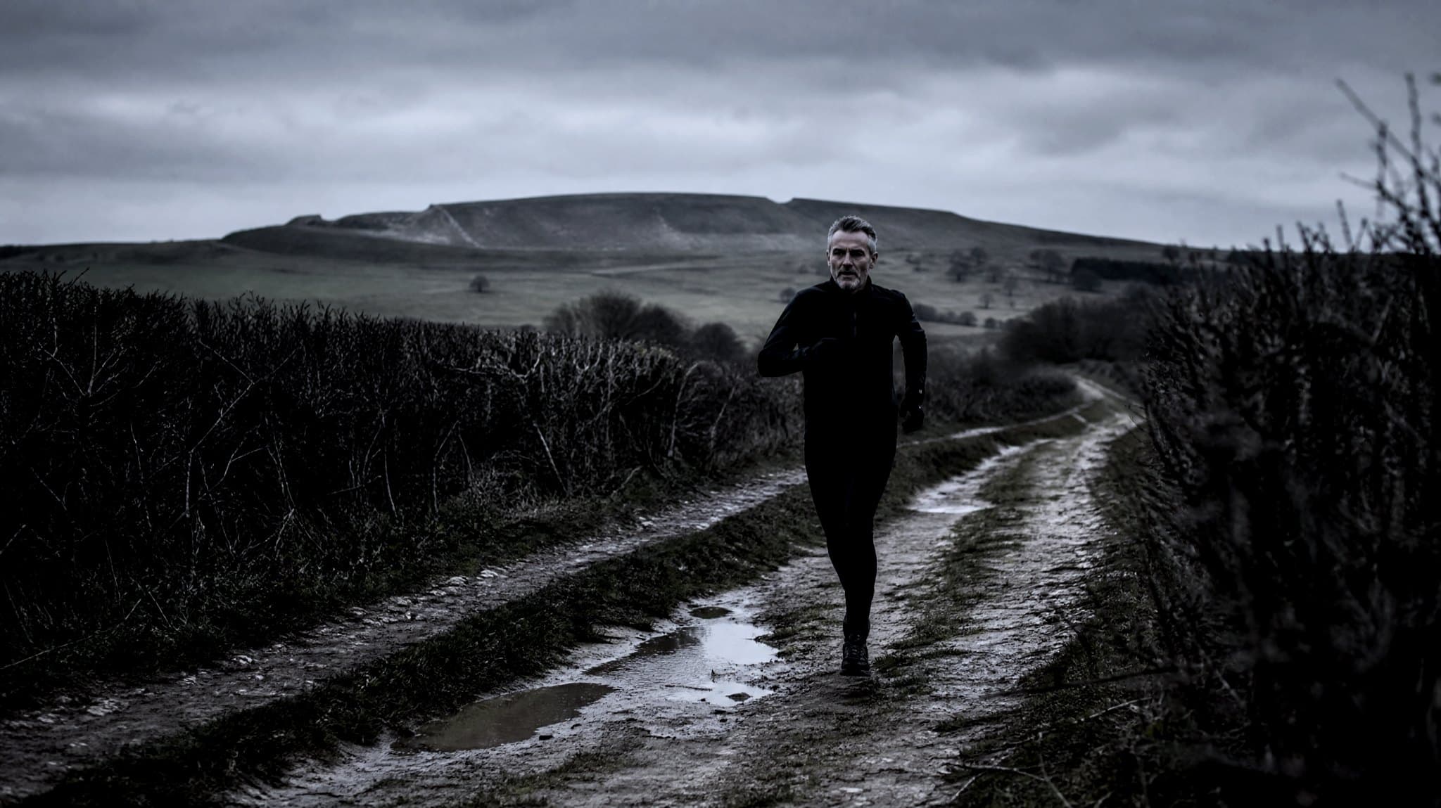 A masters-age runner on a muddy chalk track along the Ridgeway on a misty November afternoon, beech woods behind