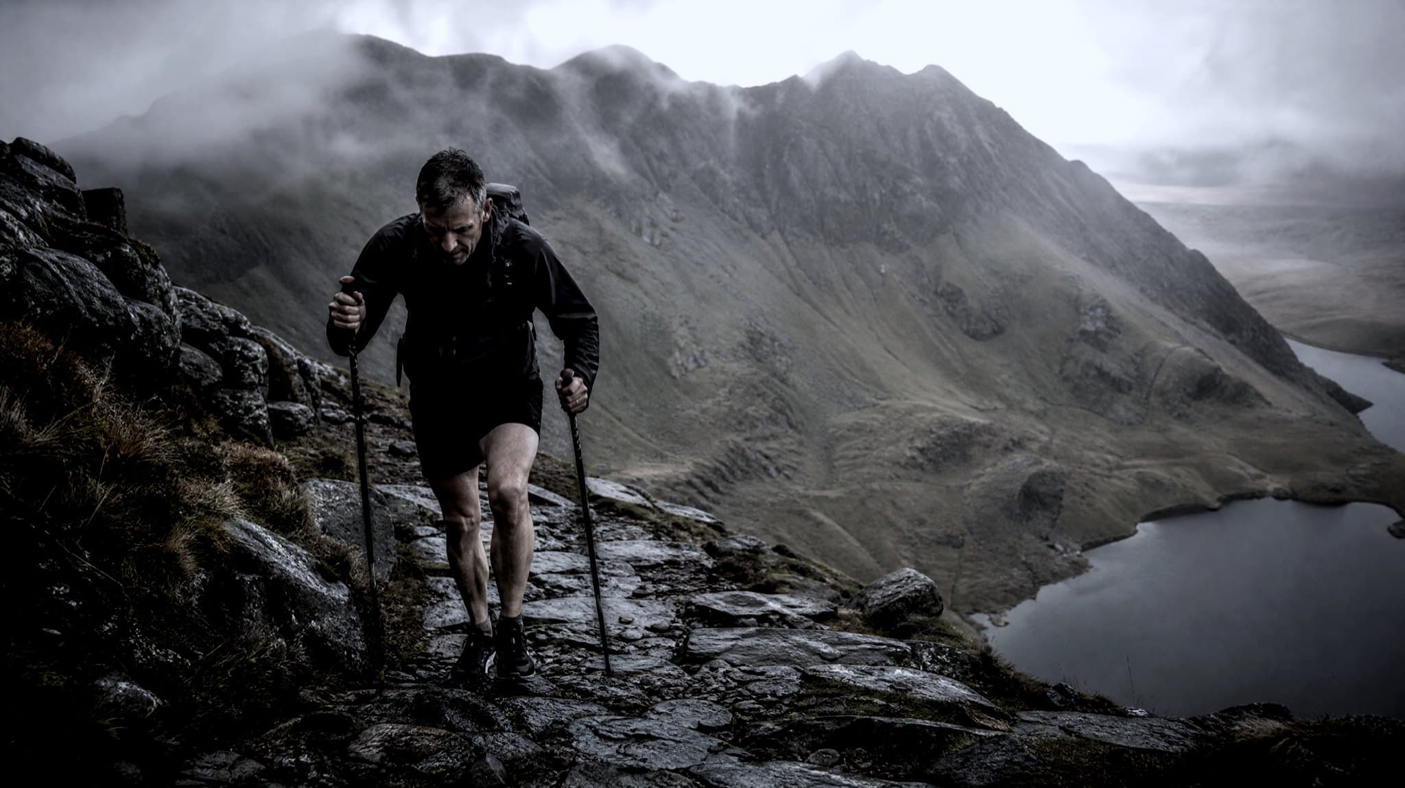 A masters-age runner with poles climbing a wet rocky path on a Welsh ridge above a lake, cloud on the summits