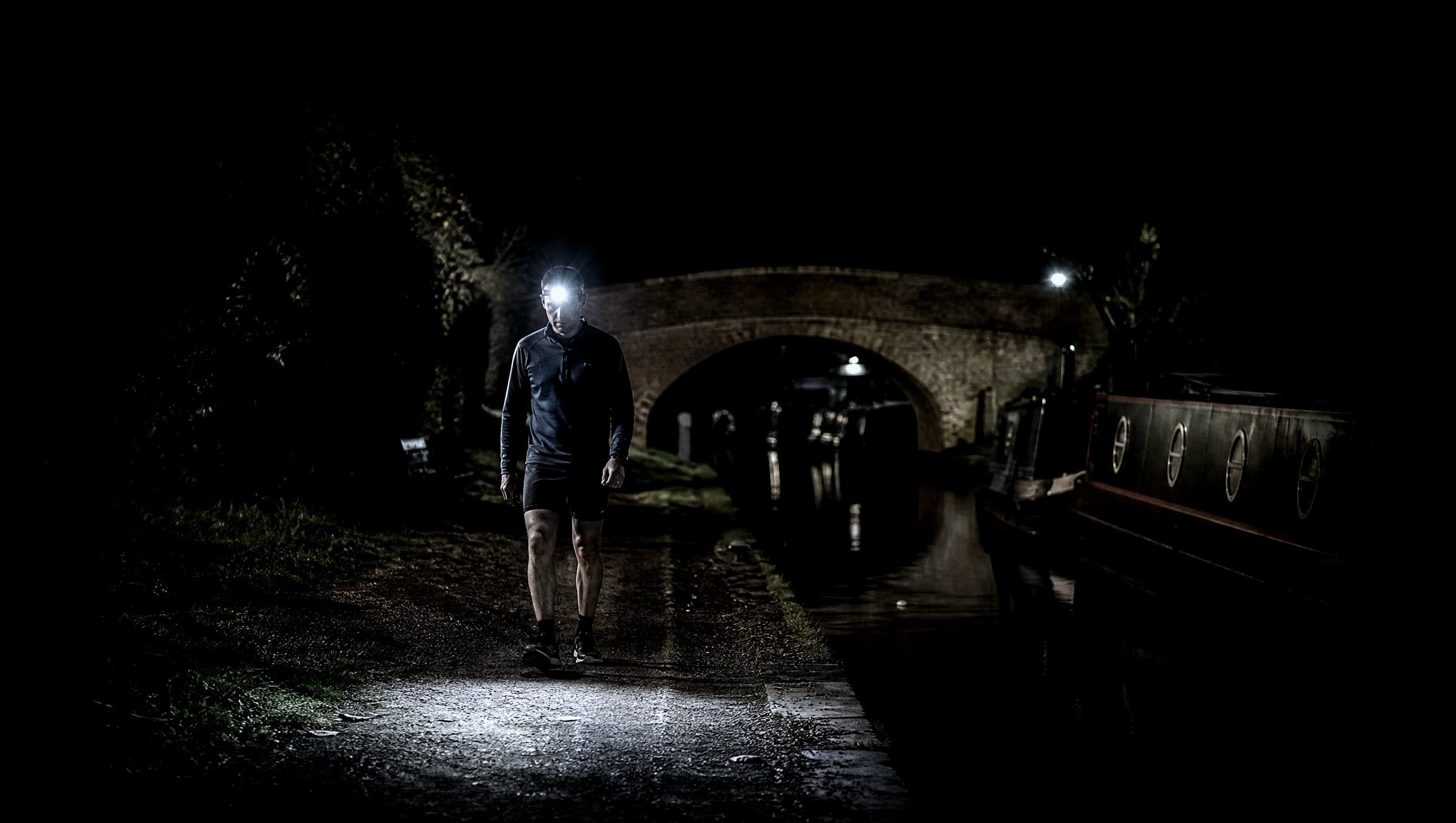 A masters-age runner walking a narrow canal towpath at night past moored narrowboats with a headtorch
