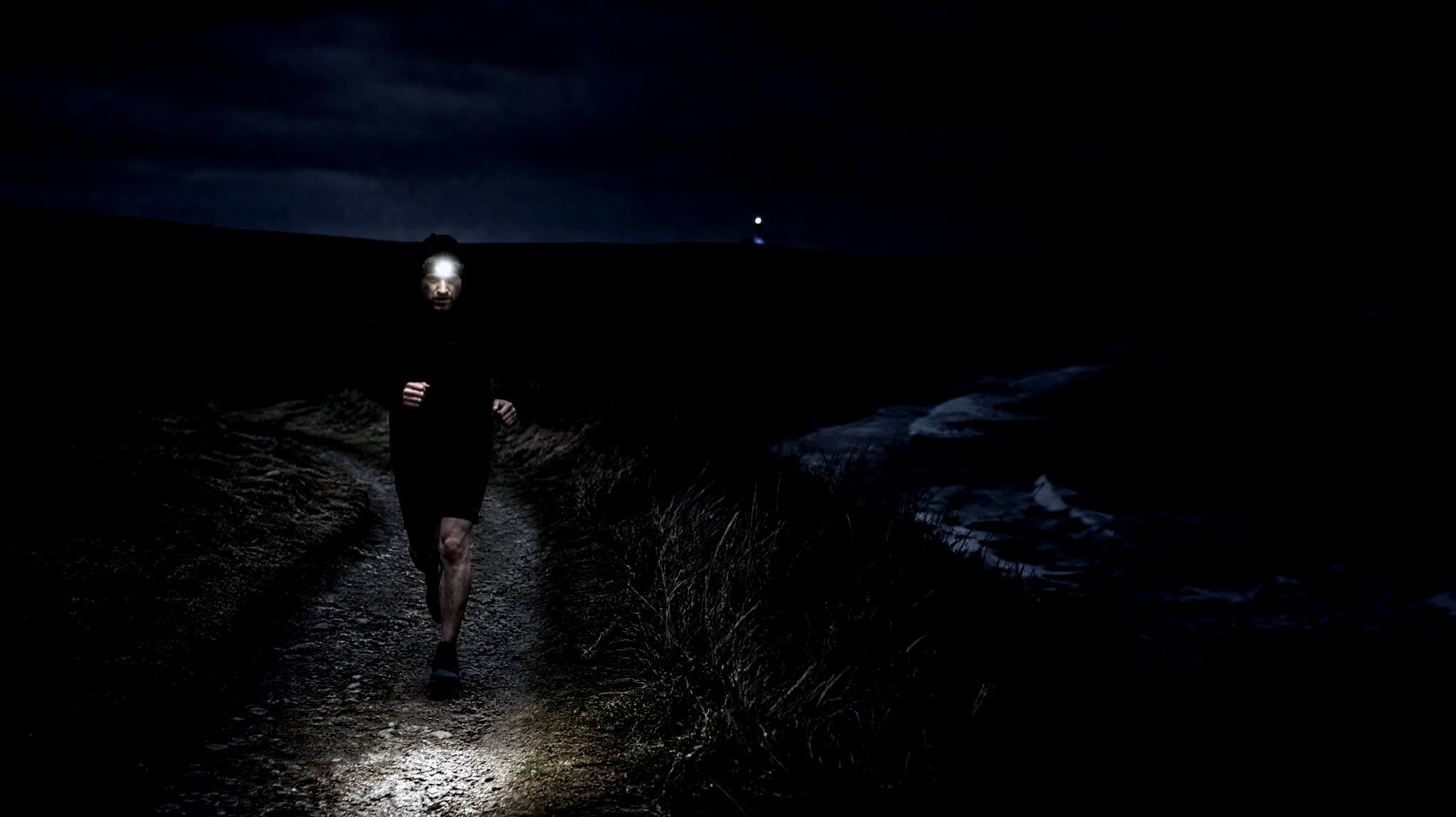 A masters-age runner with a head torch on a clifftop coastal path at night, a lighthouse on the headland behind
