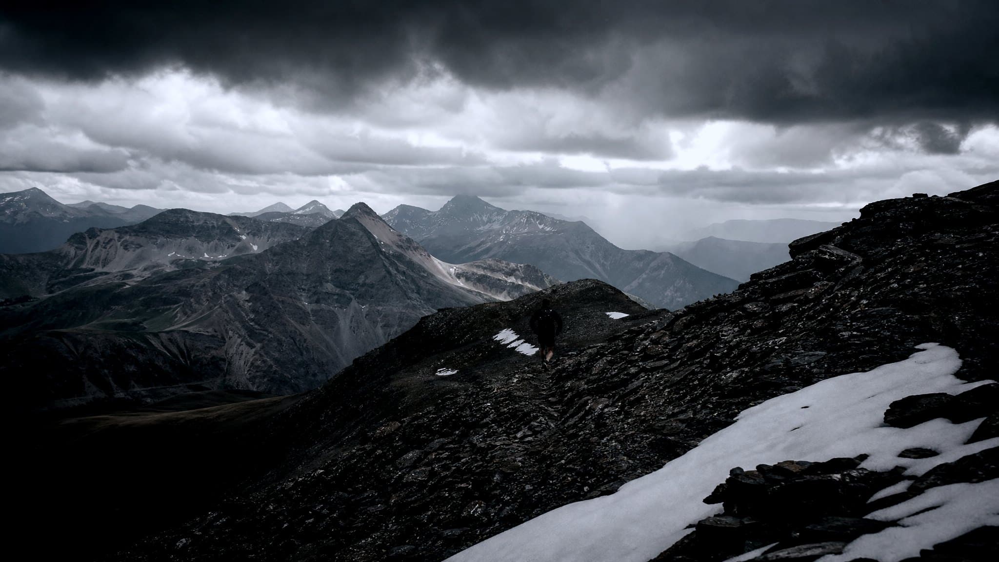 A masters-age runner hiking an alpine ridge above the treeline as storm clouds build over distant peaks