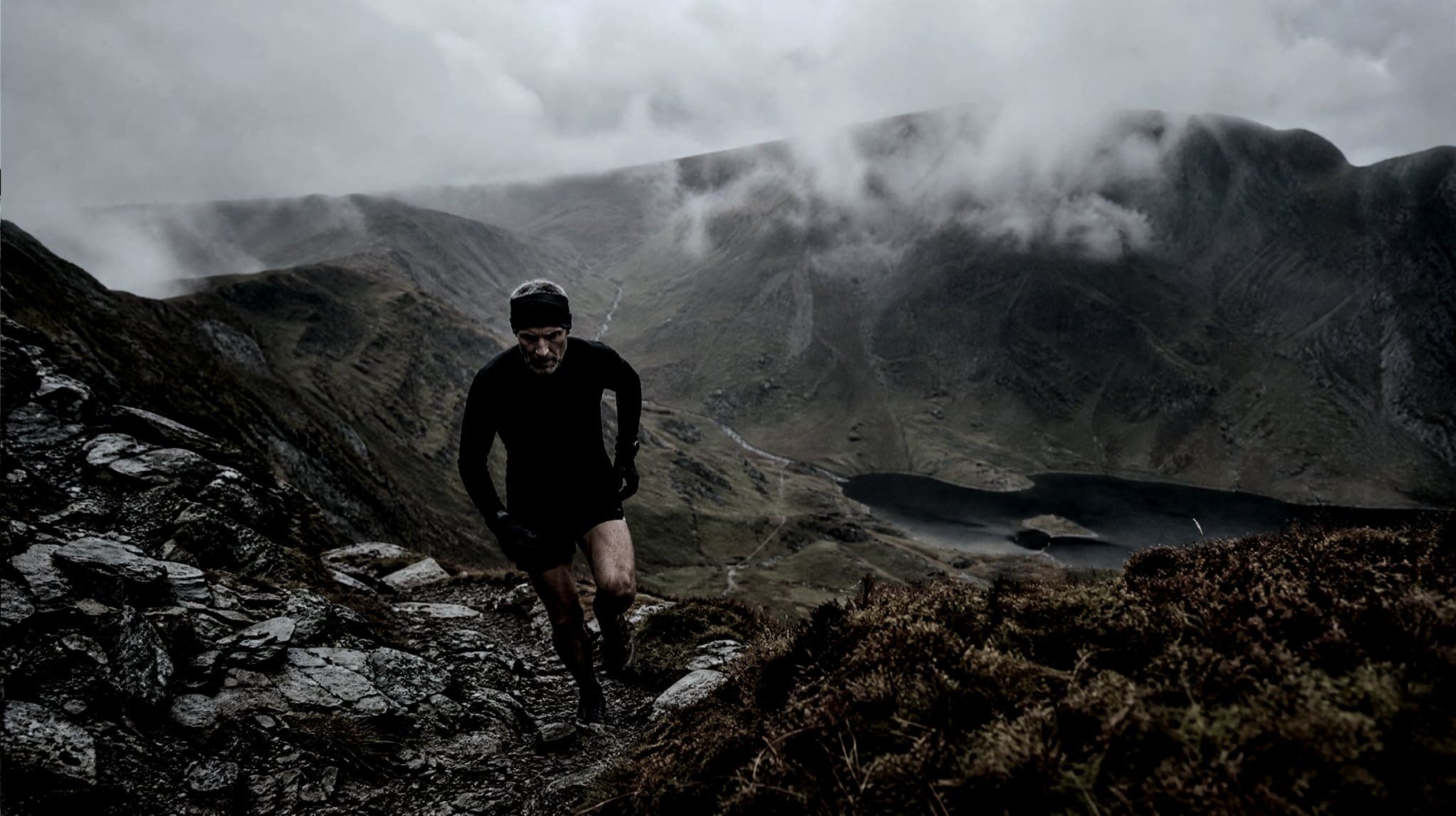 A masters-age runner picking a line down a rocky fell ridge in autumn, with a lake and low cloud below