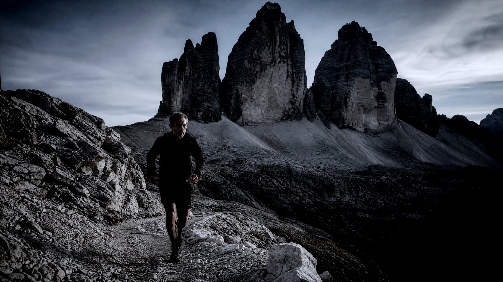A grey-haired runner on a stony path beneath the three rock towers of the Tre Cime di Lavaredo