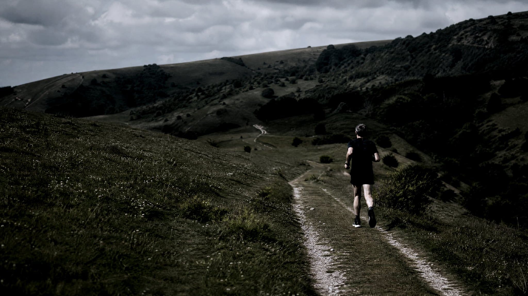 A masters-age runner climbing a chalk path onto the South Downs in early summer evening light, woodland behind