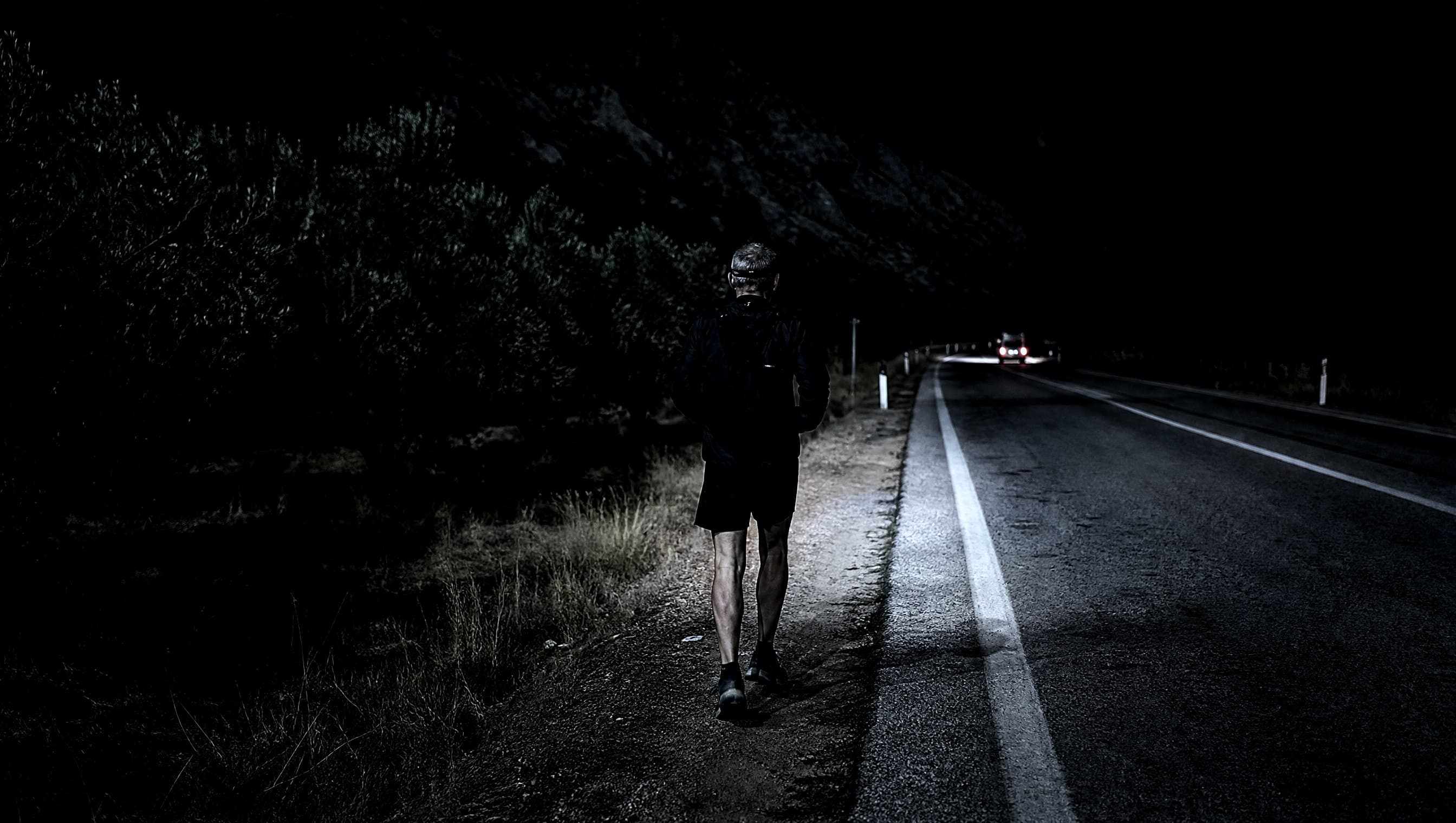 A masters-age runner alone on the shoulder of a Greek road at night with a headtorch, olive groves behind