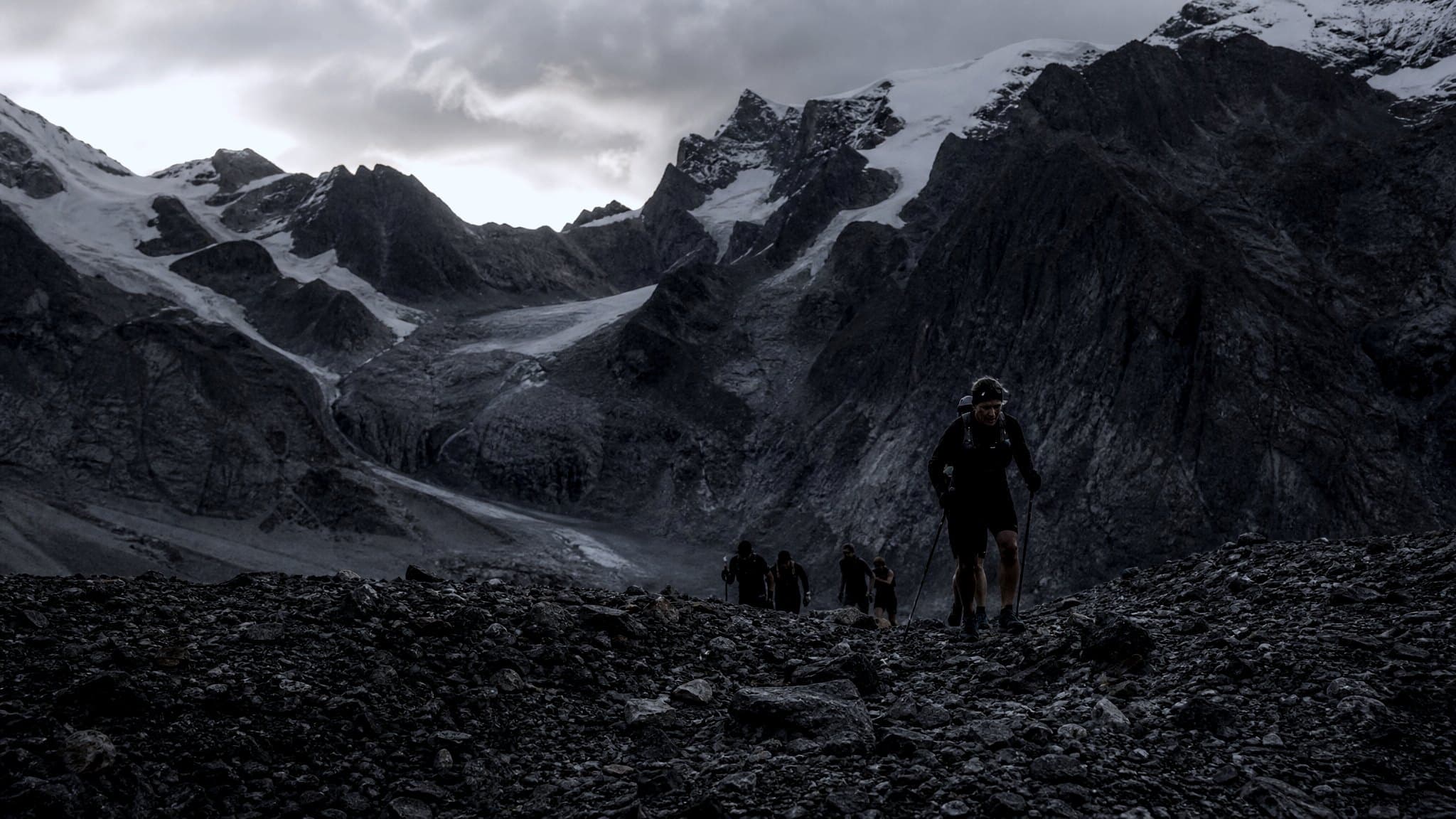 A masters-age runner with poles crossing a high stony col in the Italian Alps at dusk