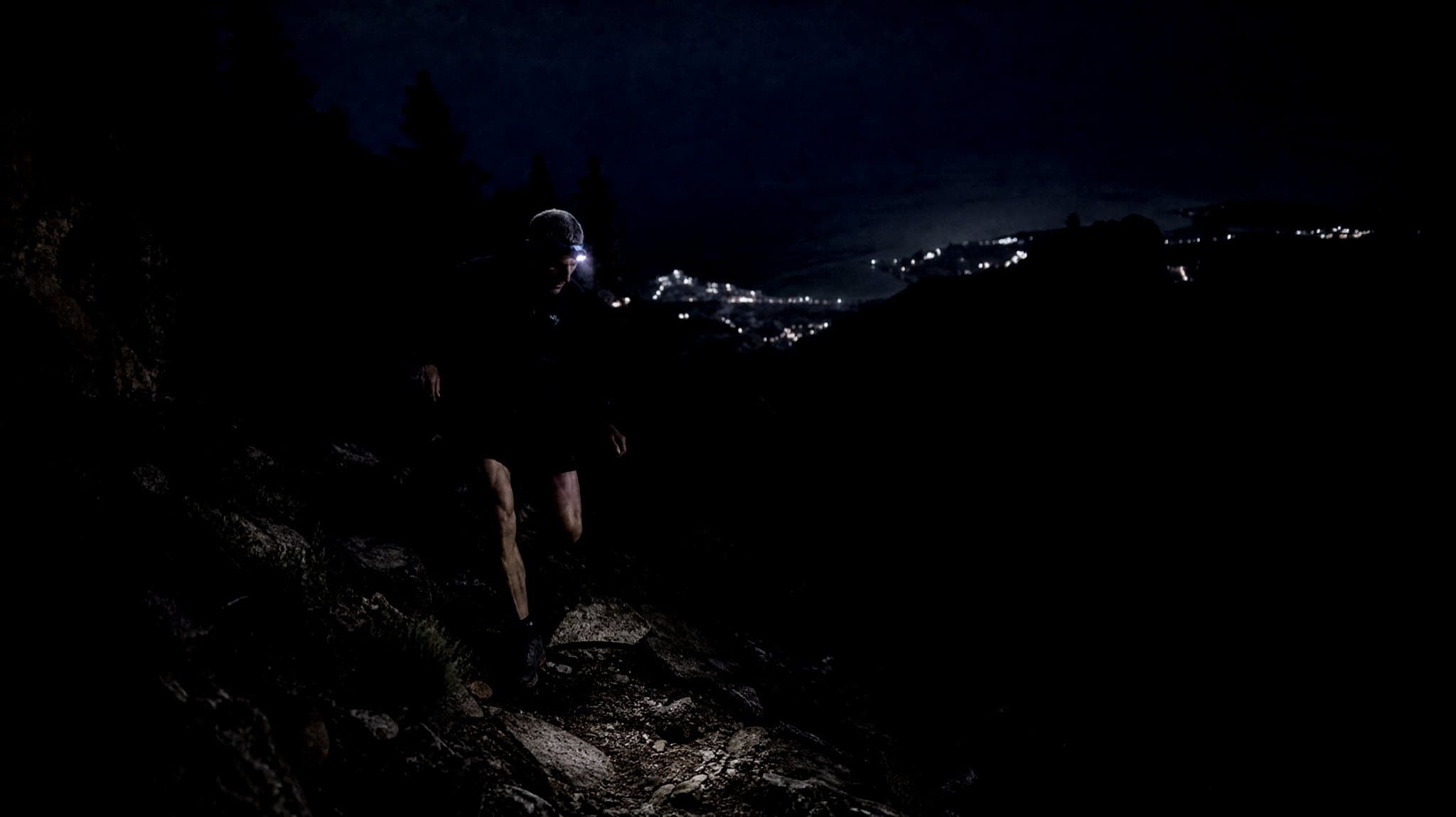 A grey-haired runner with a headtorch on a rocky mountain path at night, town lights on the coast below