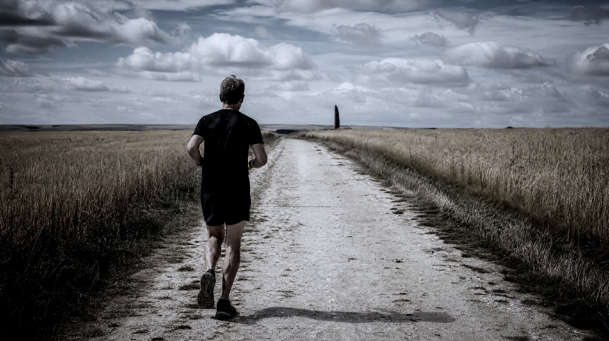 A masters-age runner on a wide chalk track along a grassy ridge in summer, with farmland falling away on both sides